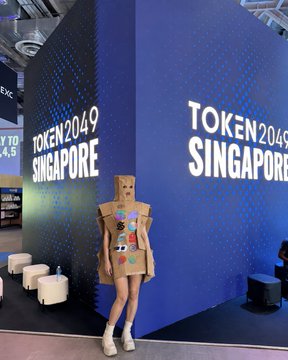 A person poses at the TOKEN2049 Singapore conference wearing a creative cardboard box robot costume adorned with colorful patches, perfectly capturing the playful "I always wanted to be different" sentiment expressed in their Day 2 recap.