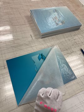 A person's gloved hand peels protective plastic film off a stack of reflective blue mirror panels resting on a wooden surface.