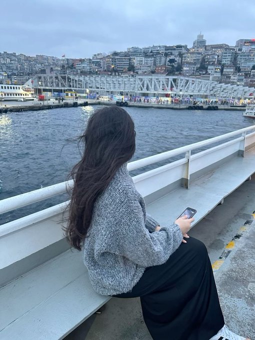 A woman with long dark hair sits on a white bench by the water, holding a phone while looking toward a cityscape and bridge.