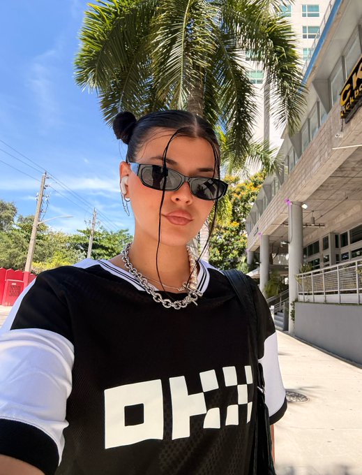 The image features a woman wearing sunglasses, a black and white t-shirt with "OKX" written across the front, and carrying a purse. The photo appears to be taken outdoors, possibly at a park or other public area. The caption of the image reads "dont ask who im rooting for. im team OKX!".