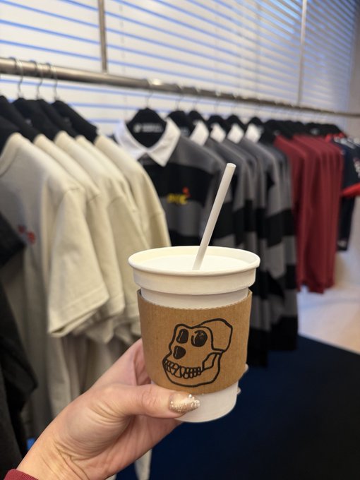 A hand holds a paper coffee cup with a cardboard sleeve featuring a skull design and a straw, with racks of clothing in the background.