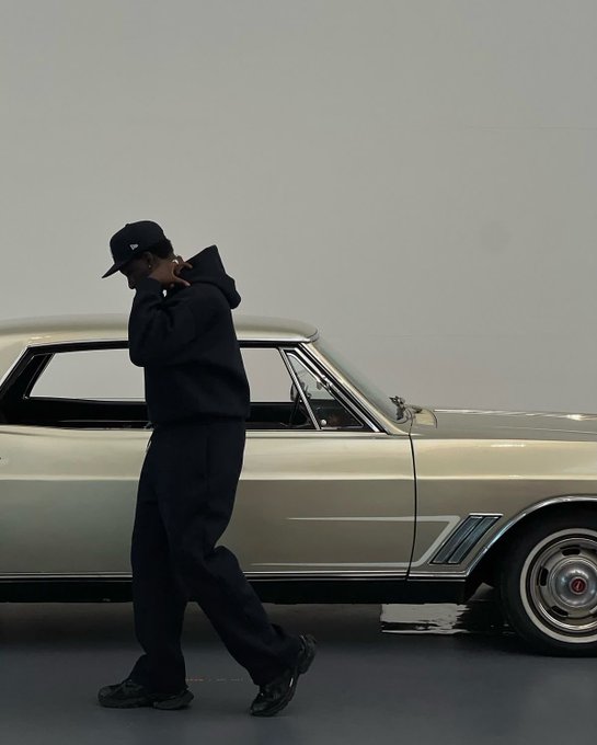 A person in all-black streetwear including a cap, hoodie, and baggy pants walks past a vintage light green car.