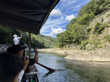 Passengers on a covered boat float down a river surrounded by lush green forested cliffs under a partly cloudy sky, with one person in a white hat standing at the front.