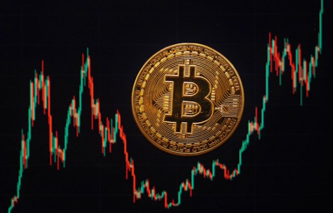 A physical Bitcoin token is prominently displayed against a backdrop of a volatile candlestick chart showing green and red price movements, symbolizing cryptocurrency's sensitivity to economic conditions. This visual ties into the post's discussion of sticky U.S. inflation at 3.4%, highlighting how persistent price pressures continue to influence both traditional markets and digital assets like Bitcoin.
