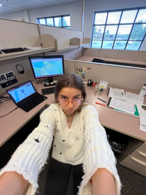 A selfie of a woman wearing glasses and a star-patterned cream sweater, sitting at an office desk with dual monitors displaying maps, with cubicles and windows in the background.