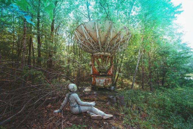 An astronaut in a spacesuit sits on the forest floor looking at a large, dilapidated radar dish in a wooded area.