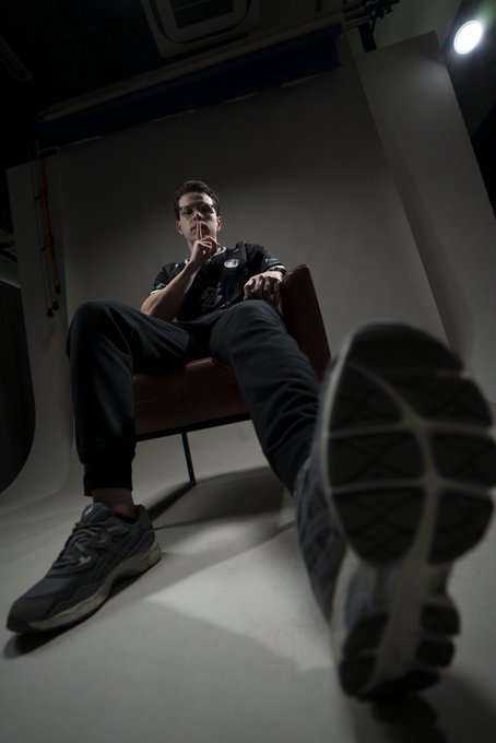 A man in a dark Nike outfit lounges confidently on a chair with his sneaker-clad foot extended toward the camera in a dramatic low-angle shot, embodying the relaxed yet ambitious energy of the post about joining a new fanbase ("@fluxogg") to "make history."