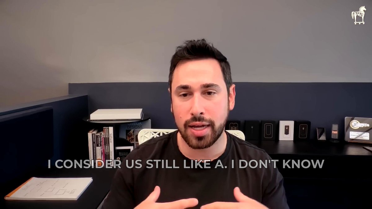 The image shows a man sitting behind a desk with his hands outstretched as if he's giving a speech or presentation. The background features a gray wall and a bookshelf filled with various items, including several books. The caption of the image reads "I consider us still like a. Don't know". This suggests that the man might be discussing the concept of Bitcoin and its principles in relation to the future of technology.