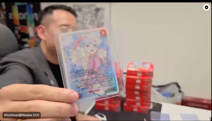 A man displays a holographic Pokémon card featuring a character with 230 HP, while holding it up in front of stacks of red booster boxes in what appears to be an office setting during a business visit. The watermark reads "Winchman@Renaiss CEO," connecting to the post about discussing card games, card-drawing machines, and asset financialization strategies with @Plus_Ultra_715.