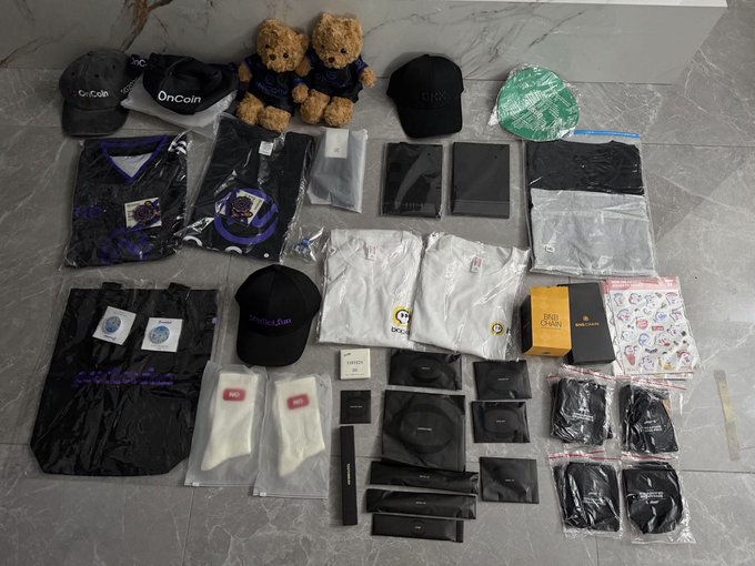 An assortment of branded merchandise and tech accessories is arranged on a marble floor, including OnCoin and BingX caps, teddy bears, t-shirts, white socks labeled 'No' and 'So', and various boxed items.