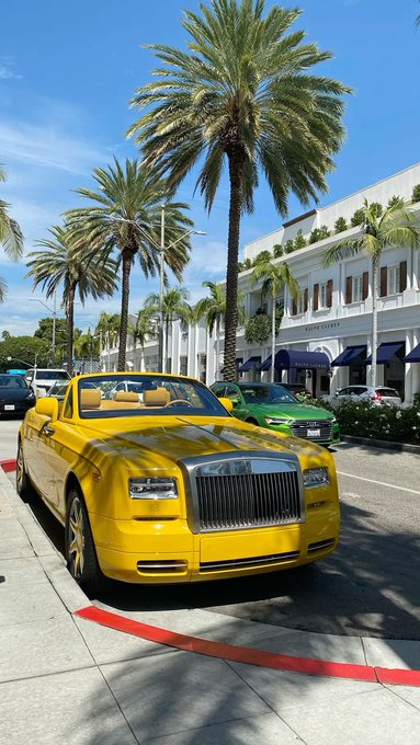 A vibrant yellow Rolls-Royce convertible parked on a street lined with tall palm trees and luxury storefronts, with a green Audi visible behind it under a bright blue sky.