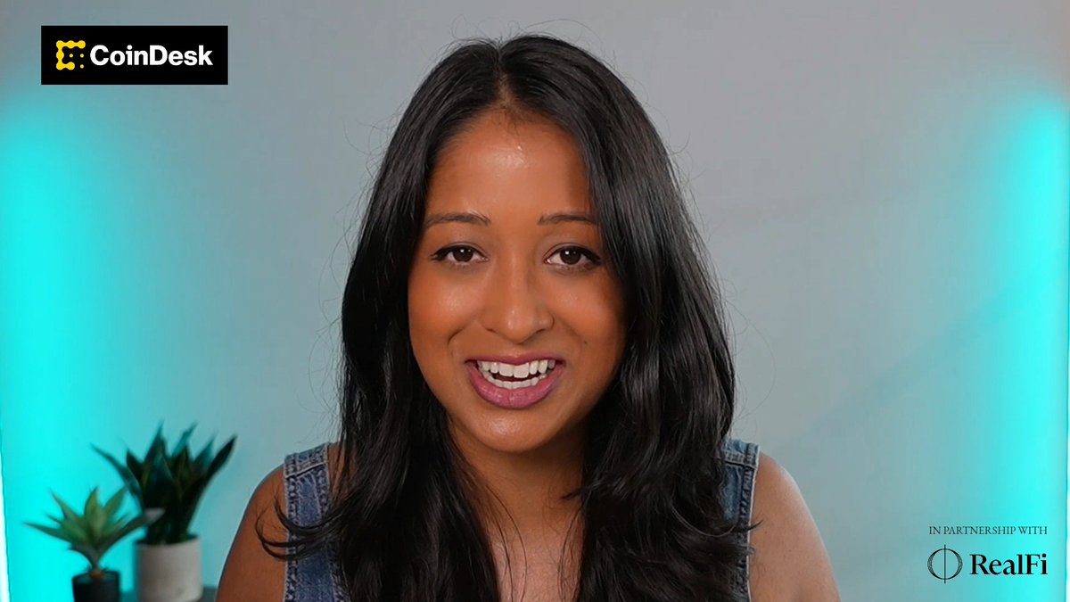 A woman with long dark hair wearing a denim top speaks to the camera in a video with a 'CoinDesk' logo in the top left and 'IN PARTNERSHIP WITH RealFi' in the bottom right.