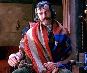A man with a mustache sits wrapped in an American flag-themed blanket in what appears to be a rustic interior setting.