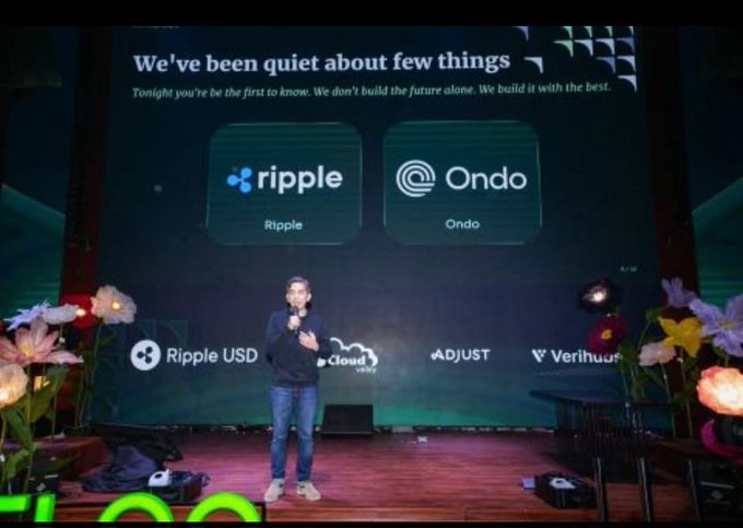 A man stands on a flower-decorated stage presenting a slide titled "We've been quiet about few things," highlighting new partnerships with Ripple and Ondo Finance, alongside logos for Ripple USD, Cloud, Adjust, and Verihubs—visually reinforcing FLOQ's announcement of its Ripple and Ondo collaboration for RLUSD and RWA tokenization in Indonesia.