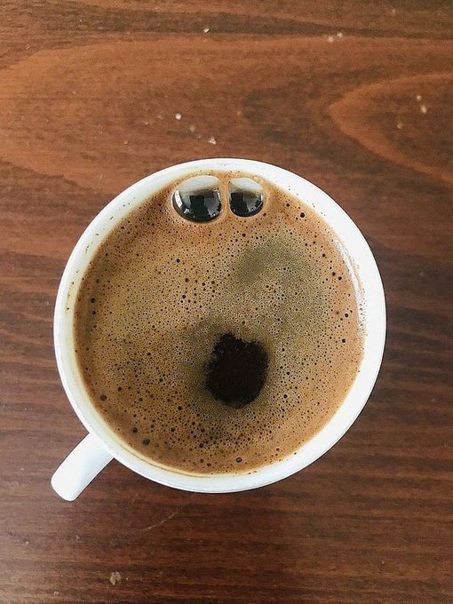 A top-down view of a white cup of coffee or espresso on a wooden table, with two coffee beans floating on the foam that resemble eyes, creating a face-like appearance.