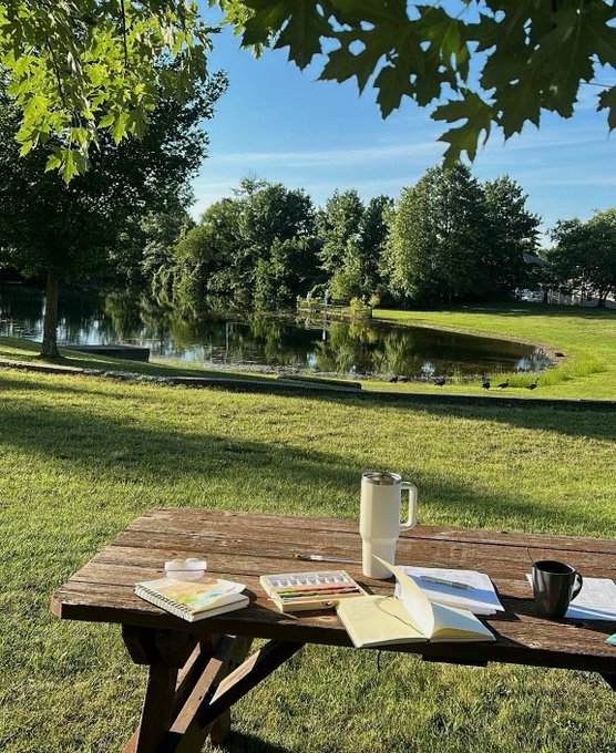 A scenic park view with a pond and ducks, featuring a wooden picnic table set up with art supplies, a thermos, and notebooks under a tree.