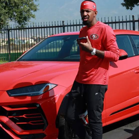 A man wearing a red San Francisco 49ers cap and long-sleeve shirt standing next to a red Lamborghini Urus SUV, holding a phone.