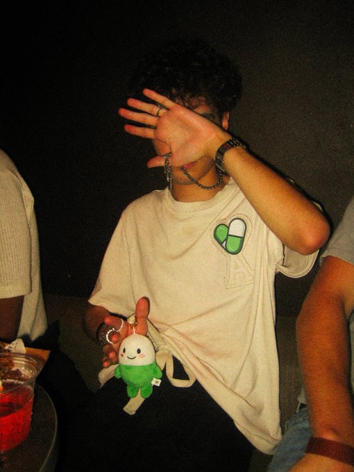 A young person at a "pump fun party" covers their face with their hand while holding a plush toy dressed in a green outfit, wearing a white t-shirt featuring a pill-shaped graphic and a silver bracelet.