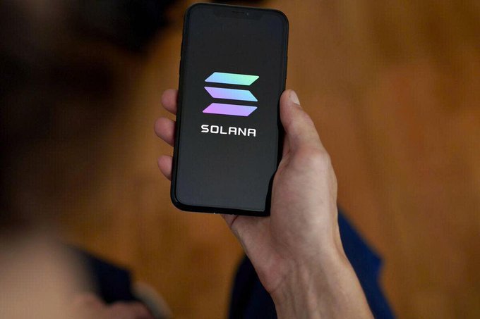A hand holds a smartphone displaying the Solana cryptocurrency logo on a black screen.