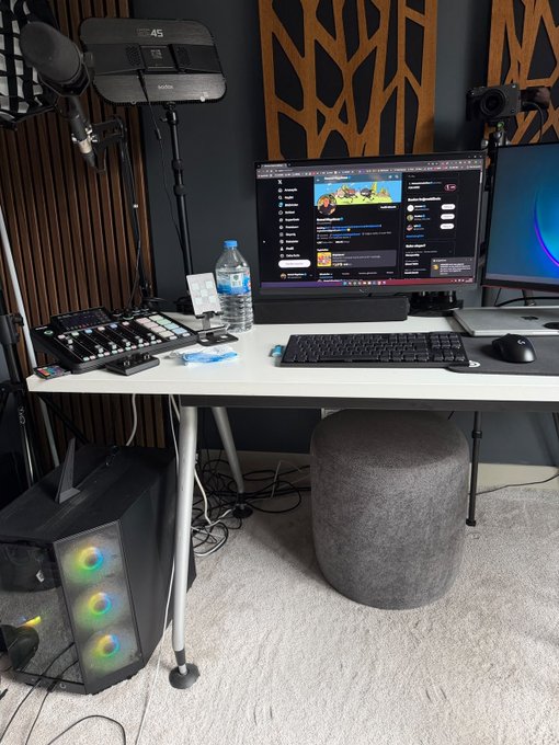 A home streaming/creator setup featuring a Godox LED panel, Rode microphone, an audio mixer, a PC tower with RGB fans, and a monitor displaying a YouTube channel. The post laments that the creator's account was hacked and people are spreading false claims, including using an outdated 2-year-old photo (likely of the Mac Mini setup) to fabricate lies about their equipment.