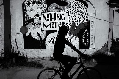 A black-and-white street photography shot of a silhouetted person riding a bicycle past a large mural reading 'NOTHING MATTERS' alongside surreal illustrations by Robert F. Mücke.
