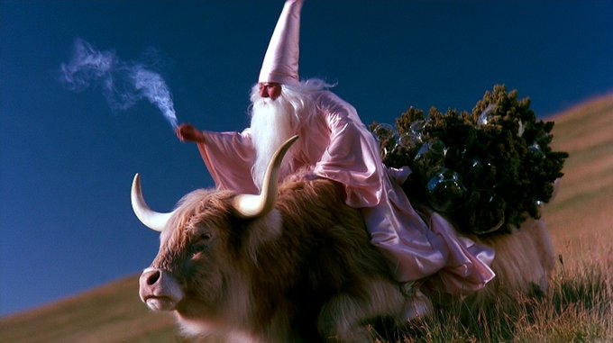 Surreal image of a wizard with a long white beard and pink robe riding a large yak through a grassy field with a Christmas tree on his back.