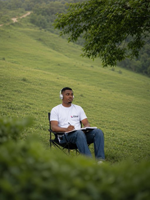 A young man wearing a white 'kleve' t-shirt, jeans, and headphones sits in a folding chair on a grassy hillside, writing in a notebook amid a lush green landscape with rolling hills and trees.