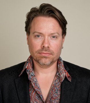 A man with brown hair and a mustache wearing a patterned paisley shirt under a dark blazer looks directly at the camera in a professional portrait.