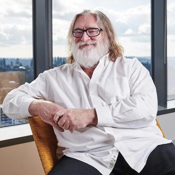 A portrait of Gabe Newell, the bearded co-founder of Valve, wearing glasses and a white button-down shirt, seated near a window.