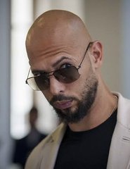 A close-up portrait of Andrew Tate wearing a cream-colored blazer over a black shirt and dark round sunglasses, with his head tilted slightly downward in a serious expression. The image accompanies a post claiming he was liquidated over 100x trading the $HYPE token with leverage, losing over $727,000, while simultaneously holding a spot position now valued at over $7 million.