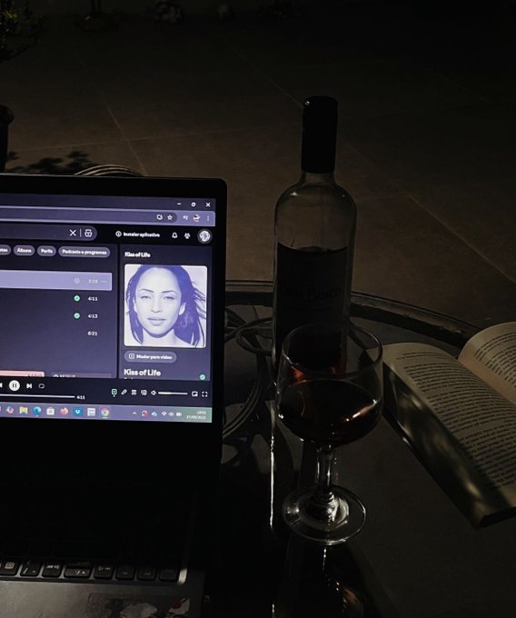 A dimly lit scene shows a laptop playing the song 'Kiss of Life' by besider reflective beside a bottle of wine and a wine glass on a glass table.