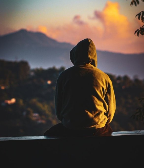 A hooded person in a yellow sweatshirt sits with their back to the camera, looking toward a softly lit mountain landscape at sunset.