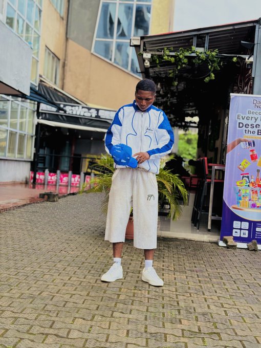 A young man wearing a blue and white jacket and beige shorts stands on a paved area, holding a blue piggy bank, with a banner reading 'Every De... the B...' visible in the background.