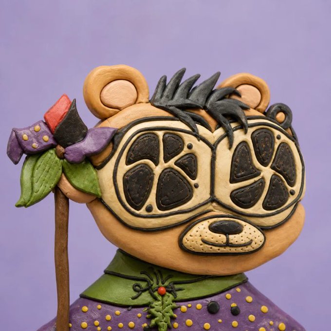 A whimsical handcrafted bear-like figurine with oversized round glasses, dark spiky hair, a purple-and-green outfit, and a flower-topped staff.