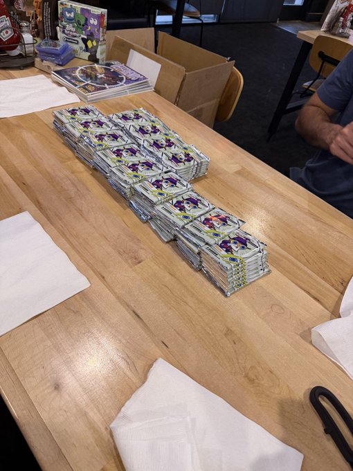 Stacks of sealed Pokemon trading card booster packs are arranged in rows on a wooden table, with a person's arm visible on the right.