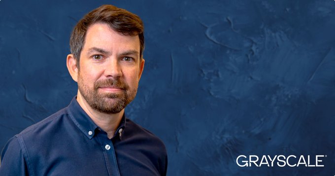 A professional headshot of a bearded man in a dark blue button-up shirt against a textured blue background, branded with the 'GRAYSCALE' logo in the bottom right corner.