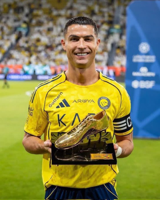 The image features a man holding up a gold trophy with his arms raised high above his head. The trophy appears to be made of metal or possibly a combination of materials such as gold and plastic. The man is wearing a yellow shirt, which contrasts nicely with the gold color of the trophy. The background shows an outdoor setting with grass, indicating that this could be a sports event or competition where the trophy was won.