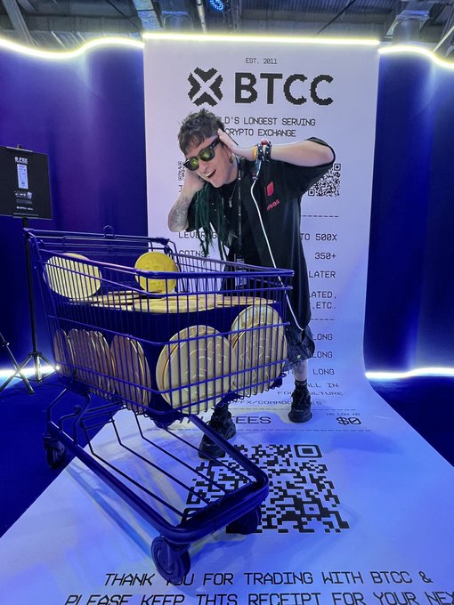A young person wearing sunglasses and a black jacket poses playfully behind a blue cart filled with large gold coins at a 'BTCC' cryptocurrency exchange booth.