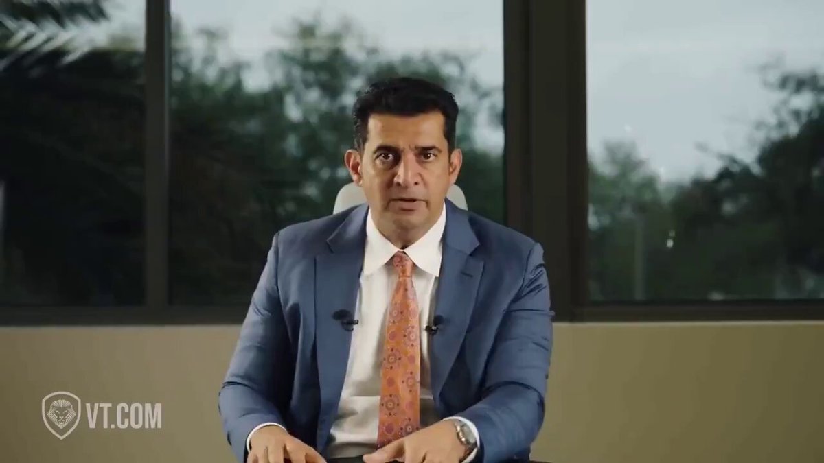 The image shows a man wearing a suit, sitting at a desk with his hands folded in front of him. He appears to be looking directly at the camera while seated in an office setting. The background features a window that provides natural light into the room. There is also text on the image that reads "Buy $XRP before SWIFT announces it." This suggests that there might be some sort of financial news or event related to cryptocurrency, and the man could potentially be discussing this topic with someone else in the office.
