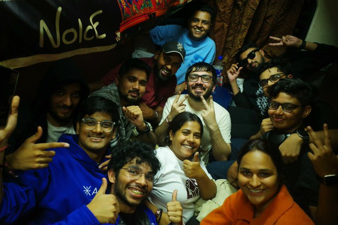 A joyful group of around ten young people pose closely together for a group photo, smiling and making hand gestures such as peace signs and thumbs-ups. In the background, a banner displays the word "NoidE," and one person wears a decorative red hat. The accompanying note about "A year since BlockTrain" suggests the photo commemorates a reunion or anniversary gathering of friends from a community or event called BlockTrain, capturing the group's enduring camaraderie and celebratory spirit one year later.