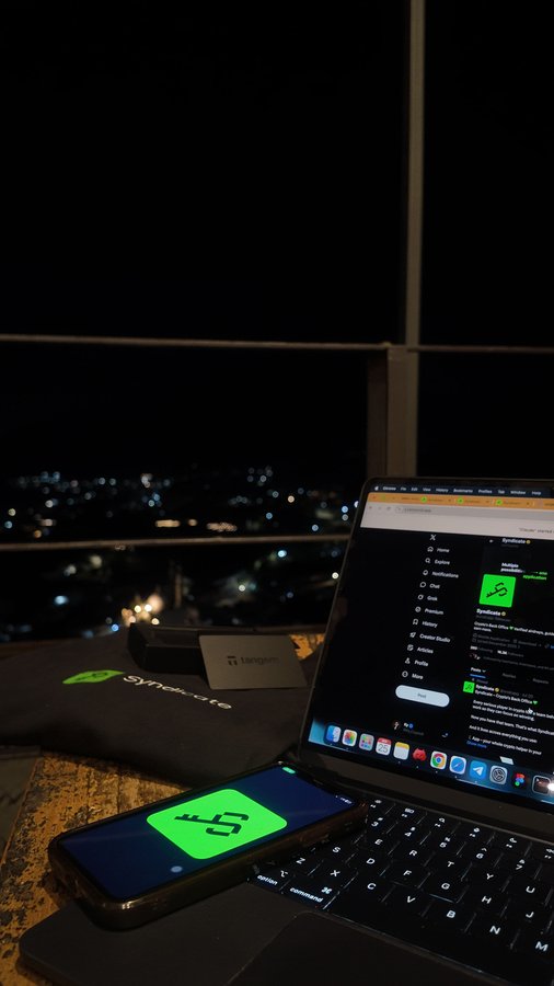 A nighttime view showing a laptop displaying a Synology interface and a smartphone showing a green '45' graphic, set against a backdrop of city lights through a window.