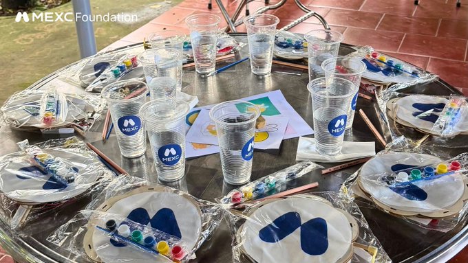 A table set up with paint supplies including cups of water, paint palettes, brushes, and paper plates with blue paint, featuring a 'MEXC Foundation' watermark and logo.