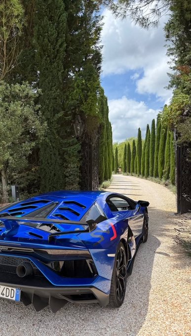 A blue Lamborghini sports car is parked on a gravel road lined with tall cypress trees under a blue sky.