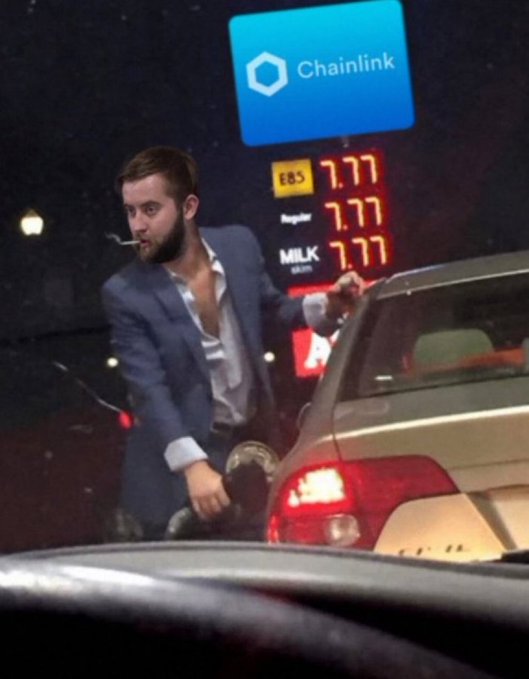 A man in a blue suit and open-collared shirt stands at a gas station at night, holding a fuel nozzle near a car with prices showing 7.77 and a 'Chainlink' sign above.