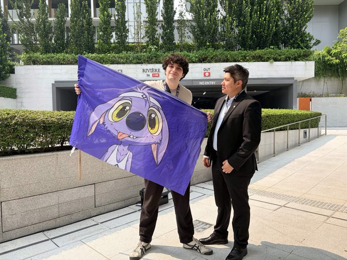 Two men stand outside a building entrance, one holding a large purple flag featuring a cartoon dog character.