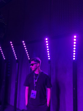 A man wearing sunglasses and a lanyard stands in a venue illuminated by purple lighting and vertical LED strips.