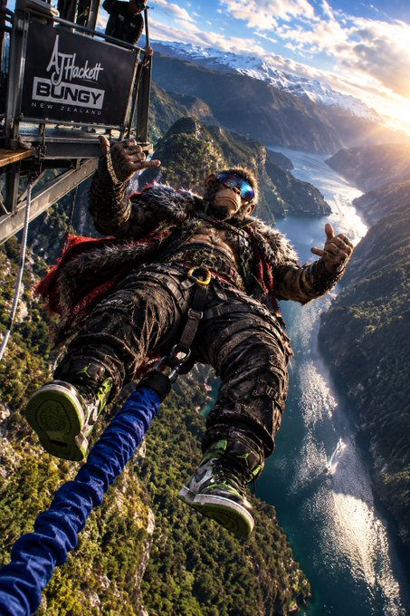 A man in a furry jacket and sunglasses is bungee jumping from the AJ Hackett Bungy platform in New Zealand, with a stunning view of the lake and mountains below.