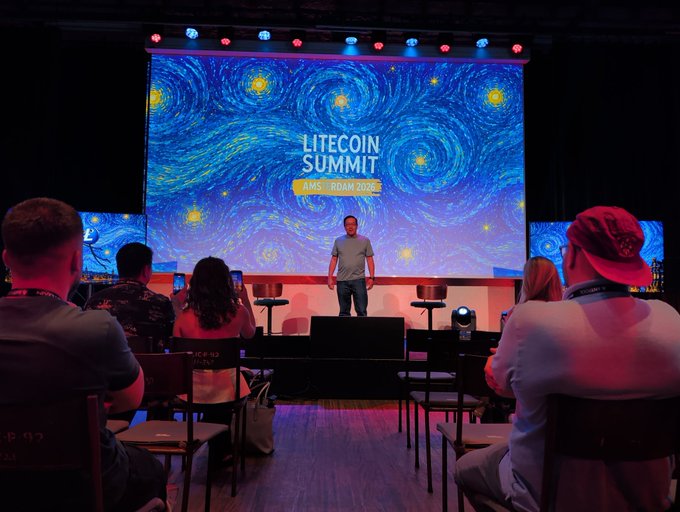 The image shows a man standing at a podium, giving a speech or presentation to an audience of people sitting in chairs around him. The event appears to be taking place in front of a large screen displaying a starry night sky with blue swirls, possibly representing Bitcoin logos and the Litecoin Summit logo. The attendees are engaged in listening to the speaker, indicating that this is likely a formal or educational event related to cryptocurrency and blockchain technology.