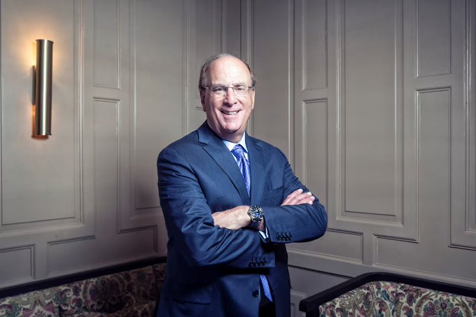 A portrait of a smiling older man with glasses, wearing a dark suit and blue tie, standing with arms crossed in an elegantly paneled room with wall sconces.
