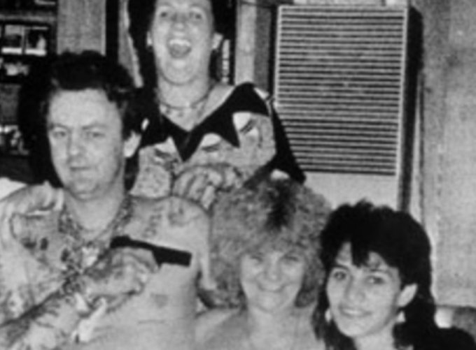 A grainy black and white photograph showing four people, including a man with a gun and a woman with an open mouth, in what appears to be an indoor setting.